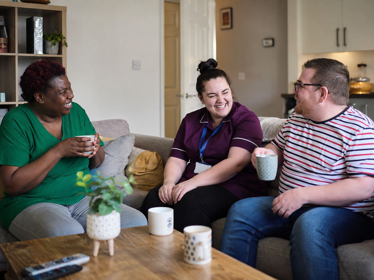 Three people laughing together on a sofa with mugs of tea
