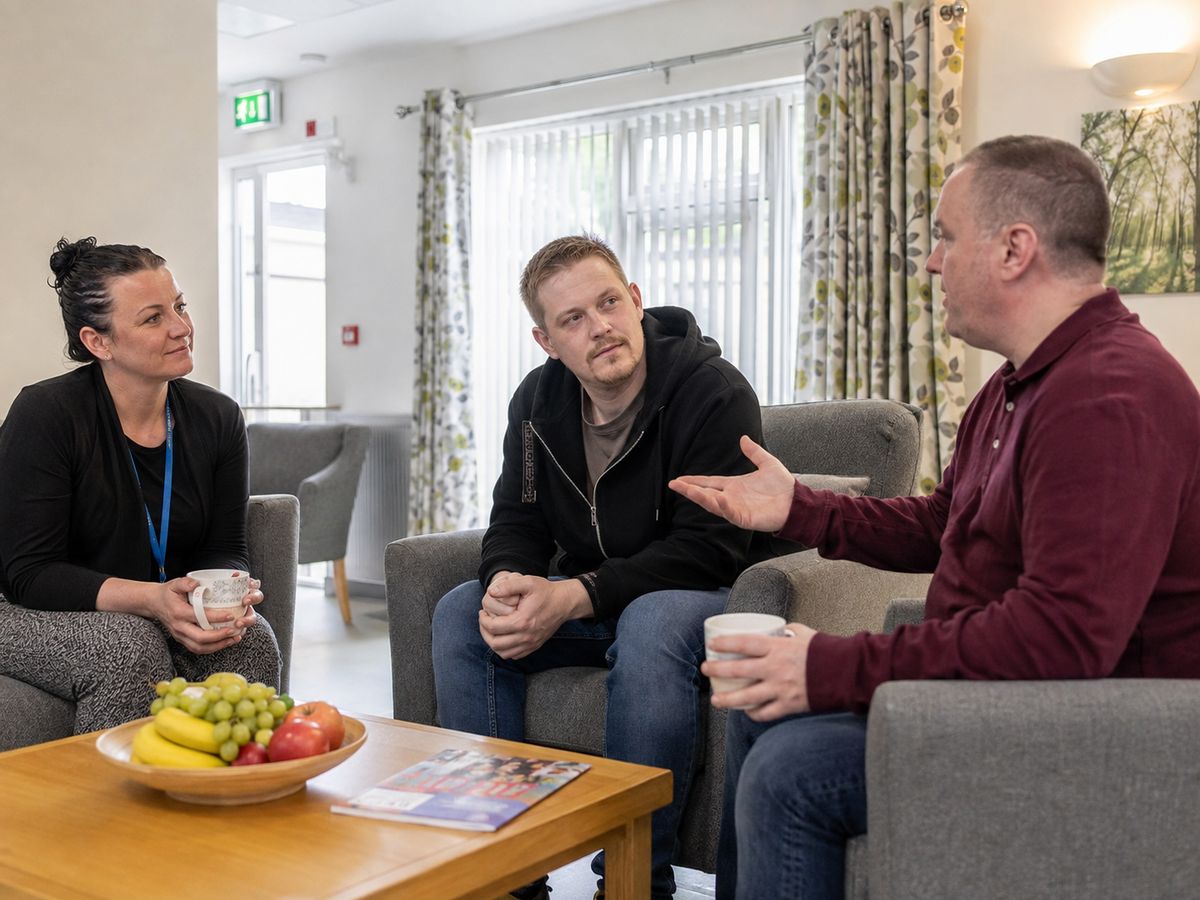 Three people, including a support worker, sitting in armchairs in a bright communal lounge